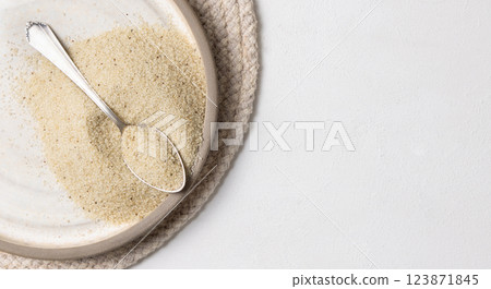 Raw uncooked fonio seeds on grey plate with a metal spoon on white table topview. African cereal Raw uncooked fonio seeds on grey plate with a metal spoon on white table topview. African cereal 123871845