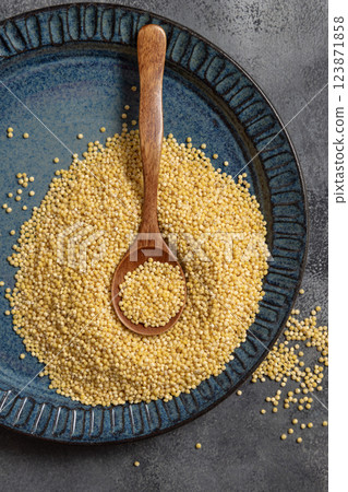 Blue plate of raw uncooked yellow hulled millet grain with wooden spoon on gray table top view Blue plate of raw uncooked yellow hulled millet grain with wooden spoon on gray table top view 123871858
