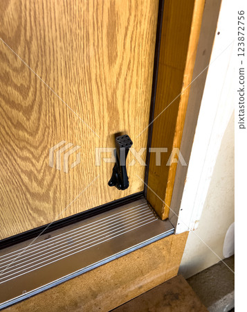 Close-up of a wooden door featuring a black metal kickstand near the bottom. The door is set within a metal threshold, and the surrounding floor has a textured surface. The kickstand is used to keep 123872756