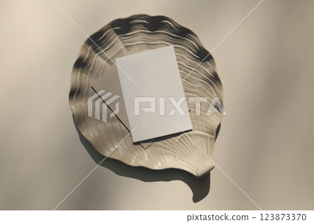 Modern summer stationery still life. Blank greeting card, invitation mock up scene with oyster shell ceramic plate. Beige table background in sunlight, long shadows. Flat lay, top view. Vacation 123873370