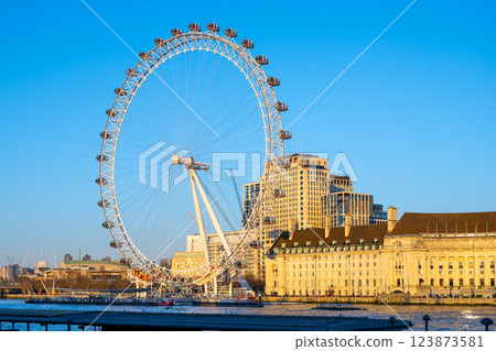 A large observation wheel stands tall against a clear blue sky, providing breathtaking views of the London skyline and the River Thames. Tourists enjoy this iconic attraction from capsule rides. A large observation wheel stands tall against a clear blue sky, providing breathtaking views of the London skyline and the River Thames. Tourists enjoy this iconic attraction from capsule rides. 123873581