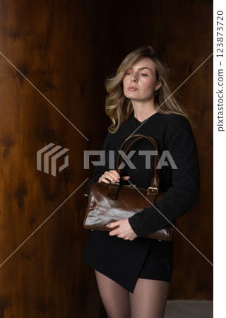A girl in a warm sweater, a black short skirt and nylon tights is posing with a brown bag. 123873720