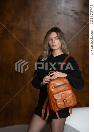 A girl in a warm sweater, a black short skirt and nylon tights is posing with a brown backpack. A girl in a warm sweater, a black short skirt and nylon tights is posing with a brown backpack. 123873741