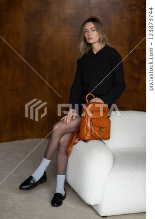 A girl in a warm sweater, a black short skirt, nylon tights, and black shoes is posing with a small brown backpack. A girl in a warm sweater, a black short skirt, nylon tights, and black shoes is posing with a small brown backpack. 123873744