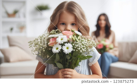 Girl holding bouquet of white and pink flowers in bright living room with blurred mother in background. Mother's Day celebration 123875273