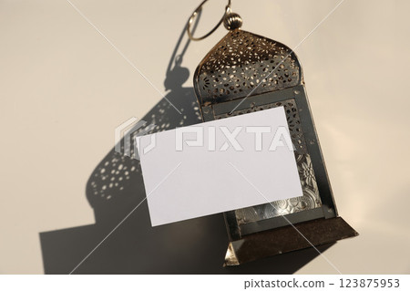 Blank greeting card, invitation template mockup on golden metal ornamental Moroccan lantern. Beige table background, shadows, top view. Festive banner still life, Muslim holiday Ramadan Kareem. Eid 123875953