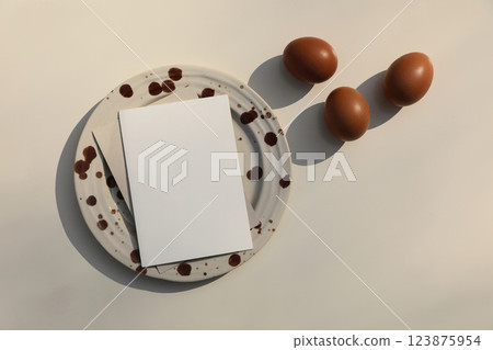 Easter still life. Brown spotted ceramic plate with blank greeting card, invitation. Hen eggs on beige table background in sunlight, long shadows. Springtime holiday home decor. Flat lay, top view Easter still life. Brown spotted ceramic plate with blank greeting card, invitation. Hen eggs on beige table background in sunlight, long shadows. Springtime holiday home decor. Flat lay, top view 123875954