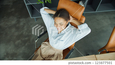 Relaxed young woman leaning back in a leather chair in a modern office, smiling with hands behind her head 123877501