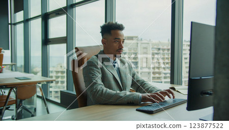 A focused businessman working on a computer in a modern office with floor-to-ceiling windows overlooking an urban cityscape, representing professionalism, technology, and workplace efficiency. 123877522