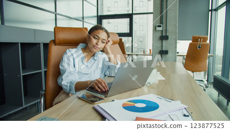 A young woman with a tired expression working on a laptop in a modern office, sitting at a desk with notebooks and large windows in the background 123877525