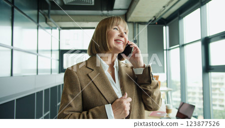 A confident mature businesswoman in a modern office talking on a smartphone, displaying professionalism, leadership, and connectivity in a contemporary corporate setting 123877526
