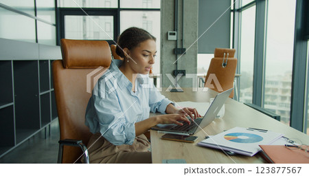 A focused young woman working on a laptop in a modern office environment, interacting with a smartphone and analyzing documents on a desk with bright natural lighting A focused young woman working on a laptop in a modern office environment, interacting with a smartphone and analyzing documents on a desk with bright natural lighting 123877567