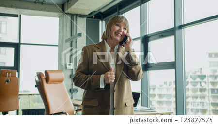 A confident mature businesswoman talking on her smartphone in a modern corporate office, with large windows and natural light, showcasing leadership and communication skills 123877575