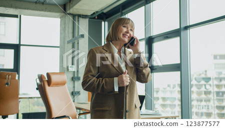 A confident mature businesswoman talking on her smartphone in a modern corporate office, with large windows and natural light, showcasing leadership and communication skills 123877577
