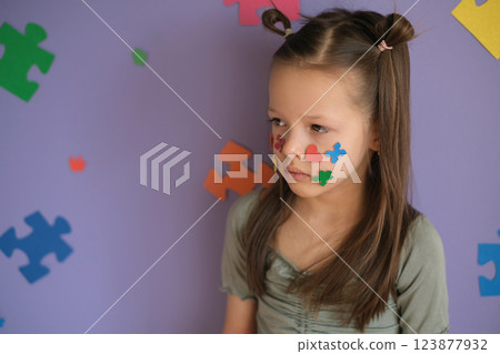 The girl looks away with a sad look. The child's cheeks are decorated with pieces of colored puzzles. World Autism Day The girl looks away with a sad look. The child's cheeks are decorated with pieces of colored puzzles. World Autism Day 123877932