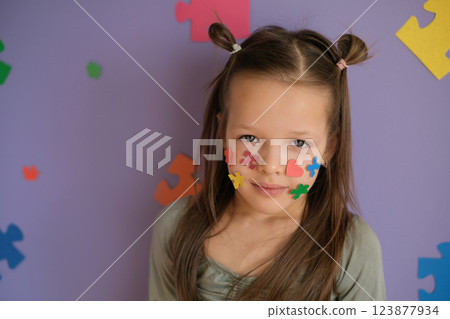 A confused, timid girl decorated her face with pieces of multicolored puzzles.  A concept photo for Autism Day. Protection and support of children with autism spectrum disorder. 123877934
