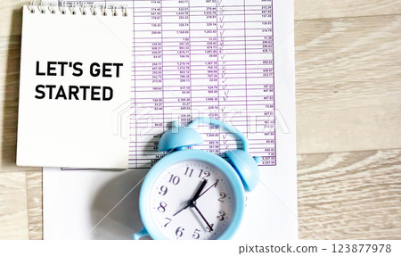 A notebook with the text "LET'S GET STARTED" on an office desk with a clock and documents.  123877978