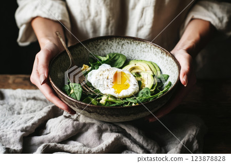 A rustic ceramic bowl filled with creamy avocado, a perfectly poached egg, and fresh spinach. The woman hands cradle the bowl with care, capturing the essence of wholesome, earthy ingredients 123878828