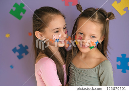Cute sisters decorated their face and purple background with pieces of multicolored puzzles. Awareness Day about autism. Difficulties in the behavior and education of children with the syndrome Cute sisters decorated their face and purple background with pieces of multicolored puzzles. Awareness Day about autism. Difficulties in the behavior and education of children with the syndrome 123879501