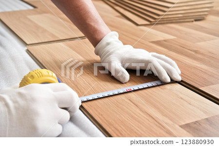 Installing laminated floor, detail on man hands in white gloves holding measuring tape over wooden tile Installing laminated floor, detail on man hands in white gloves holding measuring tape over wooden tile 123880930