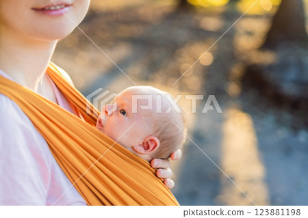 A loving mother walks through a peaceful park, carrying her baby securely in a sling. A heartwarming moment of maternal bonding, babywearing, and the joy of motherhood in nature 123881198