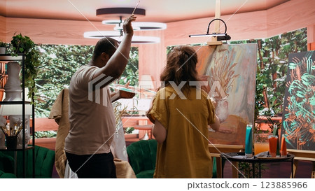 Joyful artist and her apprentice applying watercolor paint on canvas, working together to create a beautiful masterpiece in atelier. Two people painting a colorful scene with music. Camera B. 123885966