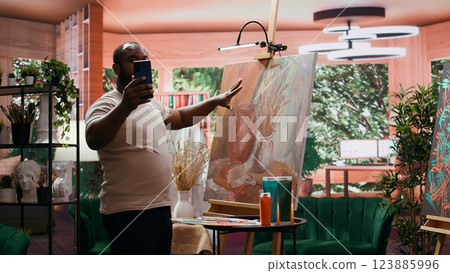 African american painter posting a picture of his masterpiece online on social media, building a better branding image for his content creation. Confident in self promotion. Camera B. 123885996
