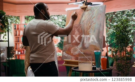 Focused man painting a new rustic artwork on canvas with brushstrokes patterns, mixing watercolors and applying to create a masterpiece. Young male artist working on his creation. Camera B. 123885998
