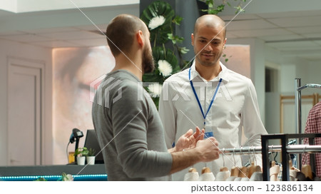 Fashion store employee assisting a male client with style choices, emphasizing customer service and presenting the new collection of quality garments. Personalized shopping experience. Camera A. Fashion store employee assisting a male client with style choices, emphasizing customer service and presenting the new collection of quality garments. Personalized shopping experience. Camera A. 123886134
