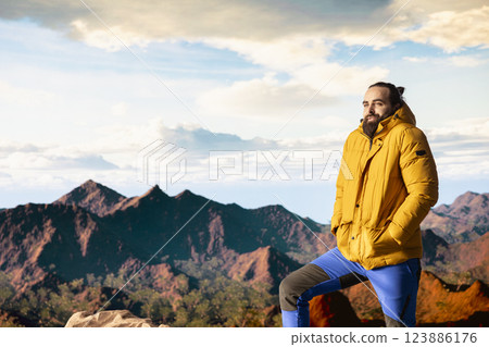Traveler stands alone at the edge of a towering mountain range, gazing at the clouds and stunning panoramic scenery. Pleased hiker showcasing personal growth and perseverance. Traveler stands alone at the edge of a towering mountain range, gazing at the clouds and stunning panoramic scenery. Pleased hiker showcasing personal growth and perseverance. 123886176