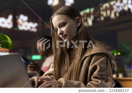Young scholar focuses intently on her exam preparations in the campus library, takes notes on her laptop from reference books in a peaceful study setting. Girl uses the general database materials. 123886237