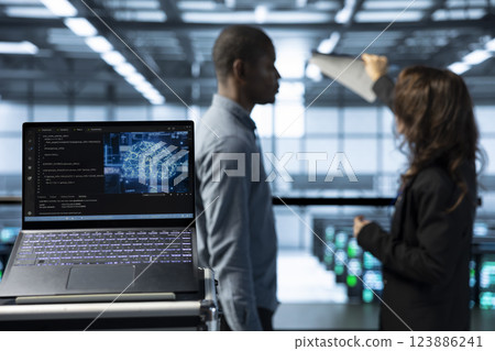 Laptop in front of data center engineers in blurry background deploying artificial intelligence tools. Close up of AI coding on notebook screen next to technicians inspecting server clusters 123886241