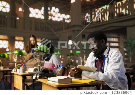 African american medic resident studying for his practice exam in the library, consulting the medical books database at institute. Young student on an internship learning for a career in healthcare. 123886964