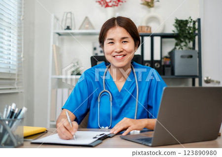 Smiling nurse in blue scrubs working at a desk with a laptop and clipboard in a modern medical office setting. 123890980