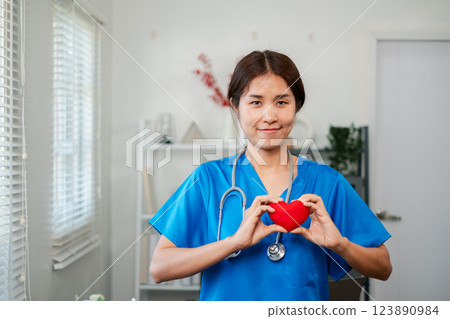 Smiling nurse in blue scrubs holds a red heart symbol, standing in a well-lit medical office with modern decor. 123890984