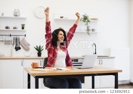 Smiling young African woman sitting at desk in modern kitchen, hands raised celebrating achievement during remote work on laptop, joyful expression 123892951