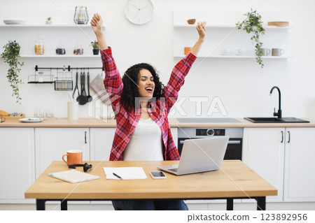 African woman in casual clothes raising arms joyfully at desk in kitchen workspace. Expressing triumph and cheer. Modern home office setup with laptop, smartphone, and notebook. 123892956