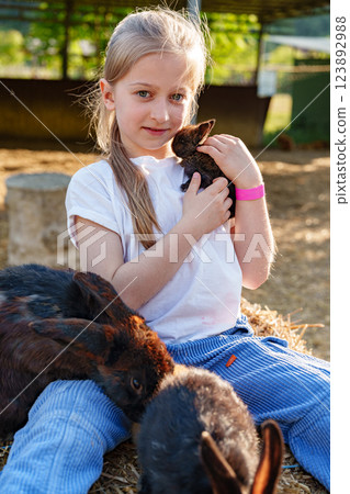 Young girl holds a small rabbit while sitting among playful bunnies at a farm during a sunny afternoon Young girl holds a small rabbit while sitting among playful bunnies at a farm during a sunny afternoon 123892988