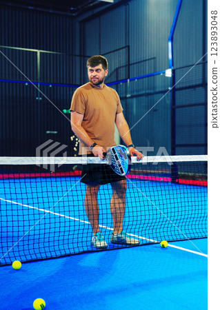 Male player preparing for a paddle tennis match on a vibrant blue court with balls scattered around 123893048
