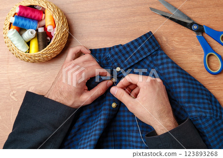 A person sewing a button on a blue plaid shirt with sewing tools and threads on a wooden table A person sewing a button on a blue plaid shirt with sewing tools and threads on a wooden table 123893268