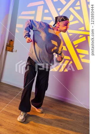 A man in a grey sweatshirt and black pants dances in a studio with a colorful light projection on the wall A man in a grey sweatshirt and black pants dances in a studio with a colorful light projection on the wall 123893444