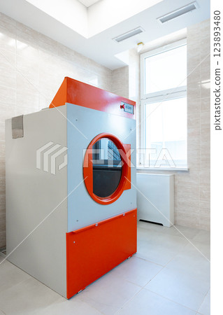 Modern laundry room featuring a large industrial washer with orange and gray design in a well-lit environment Modern laundry room featuring a large industrial washer with orange and gray design in a well-lit environment 123893480