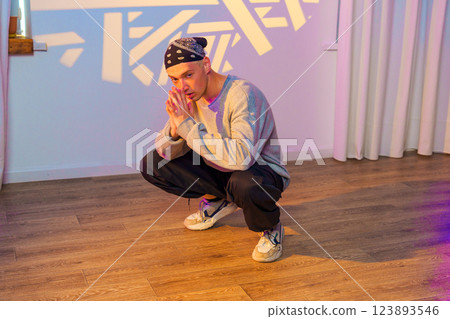 A young man wearing a grey sweatshirt and black pants sits in a squat position on a wooden floor 123893546