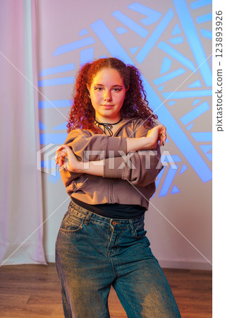 A young woman with curly red hair stands in a studio, posing with her arms crossed. She is wearing a light brown hoodie and blue jeans 123893926