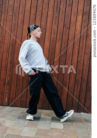 A young man wearing a grey sweater, black pants, and a bandana walks past a wooden wall A young man wearing a grey sweater, black pants, and a bandana walks past a wooden wall 123894076