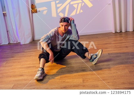 A young man sits on a wood floor in a dance studio, wearing a bandana and sneakers 123894175