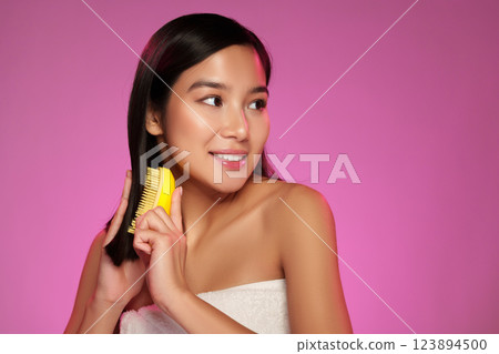 Woman grooming her hair with a yellow comb in front of a vibrant pink background during a beauty routine 123894500