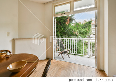 A modern dining area featuring a wooden table and bowls, leading to a balcony with greenery visible outside. Light floods in through large windows. A modern dining area featuring a wooden table and bowls, leading to a balcony with greenery visible outside. Light floods in through large windows. 123894705