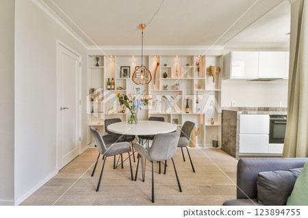 A stylish dining area featuring a round table surrounded by grey chairs, enhanced by warm lighting and decorative shelves showcasing plants and accessories. 123894755