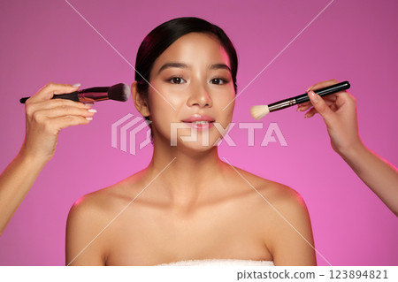 Two artists applying makeup on a model with pink background during a beauty session in a studio setting 123894821